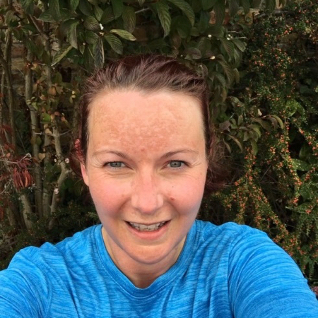 Selfie head and shoulders photo of a white woman wearing a blue running t shirt and smiling at the camera. A tall hedge fills the background. 